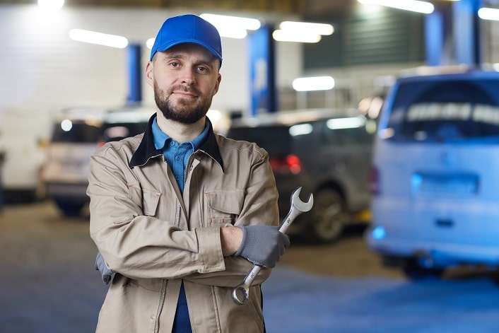 Horizontal waist up portrait of mature Caucasian auto mechanic standing with arms crossed holding spanner looking at camera, copy space
